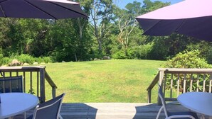 Eine Terrasse mit zwei weißen Tischen und Stühlen, beschattet von violetten Sonnenschirmen, mit Blick auf eine grüne Wiese mit Bäumen.