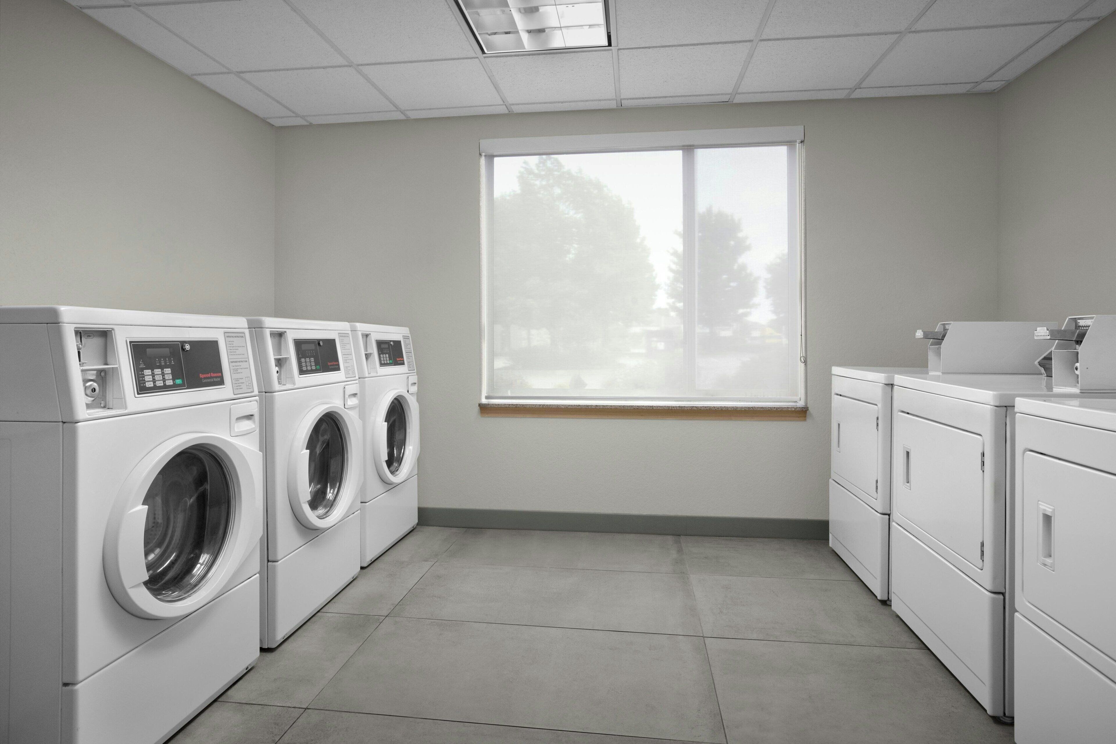 A laundry room with four washing machines and a dryer, a window with a view of trees, and a tiled floor.