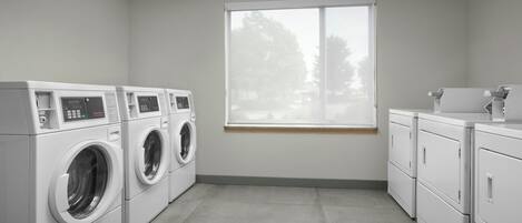 A laundry room with four washing machines and a dryer, a window with a view of trees, and a tiled floor.