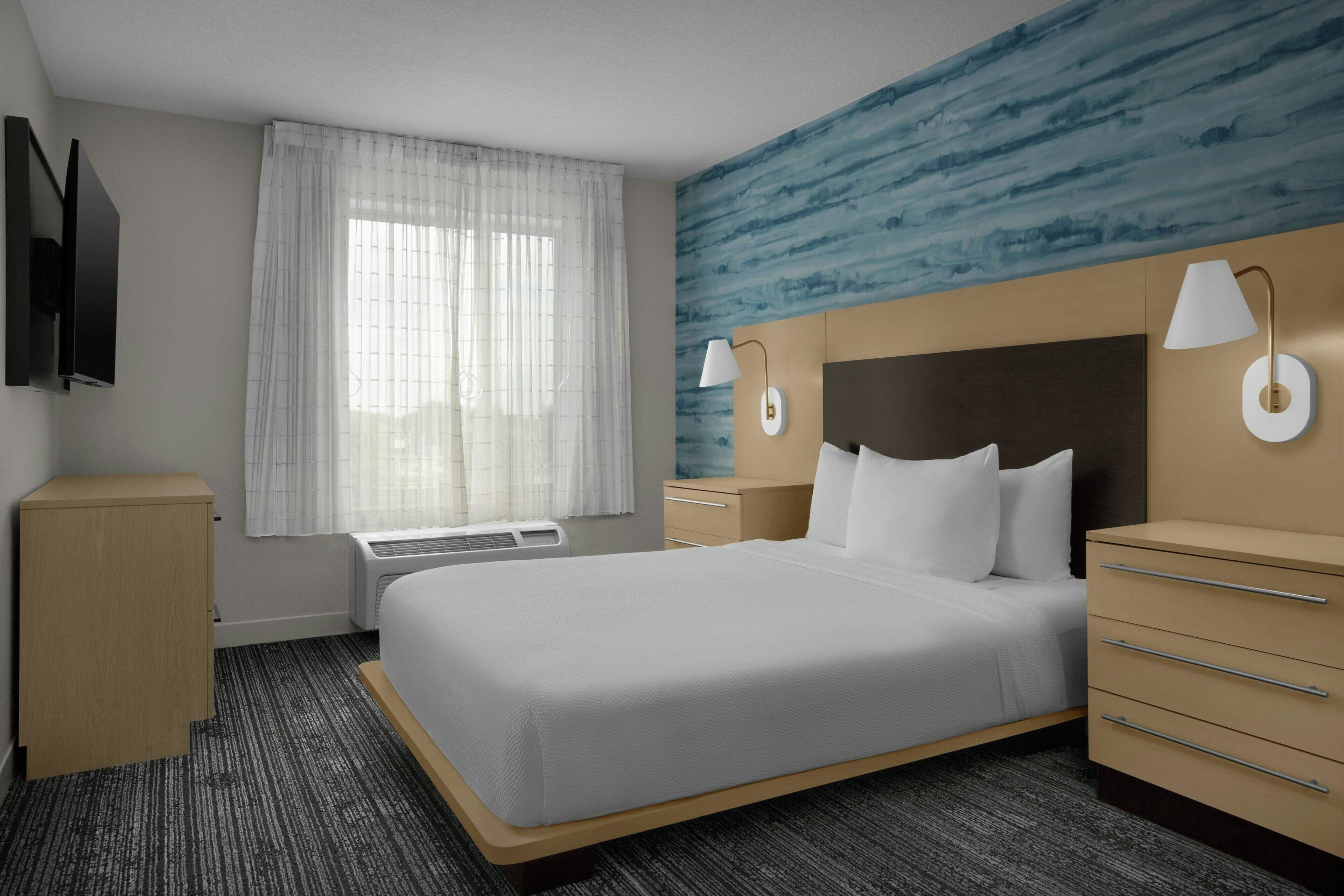 A hotel room with a bed, a desk, a dresser, a TV, and a window with curtains.