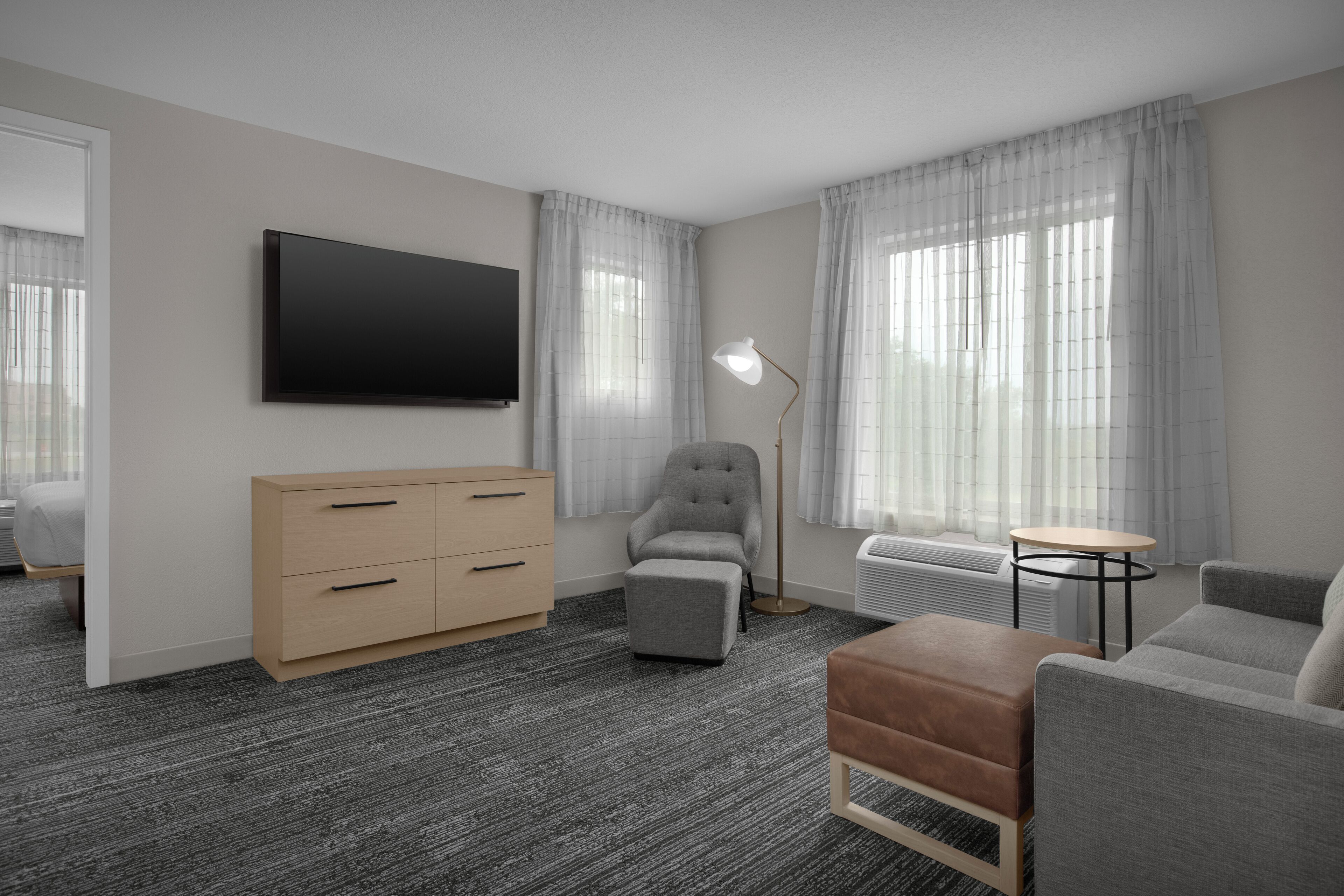 Une chambre d’hôtel moderne équipée d’un téléviseur à écran plat, d’une commode en bois, d’un fauteuil gris, d’un pouf marron, d’une petite table ronde et d’un lampadaire.
