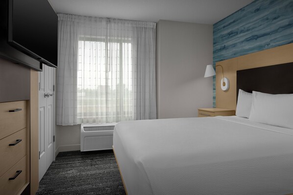 Une chambre d’hôtel comprenant un lit, un téléviseur à écran plat, une fenêtre avec des rideaux légers, une lampe fixée au mur et un mur orné d’un motif bleu.