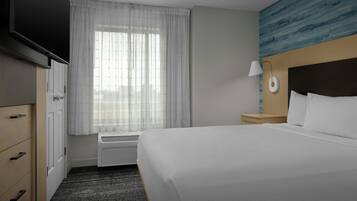 A hotel room with a bed, a TV, a window with curtains, a nightstand with a lamp, and a wall with a blue pattern.