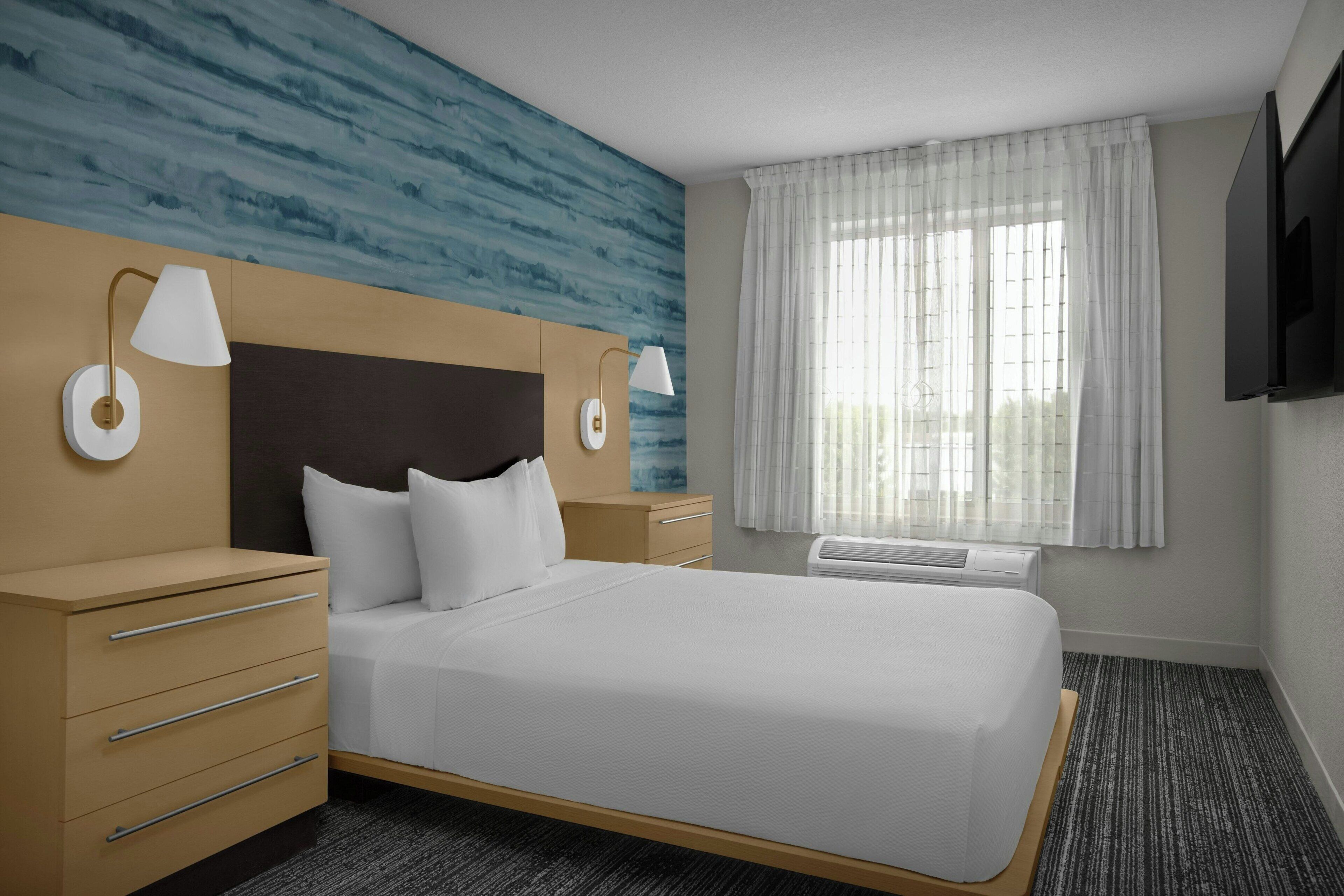 A hotel room with a bed, bedside tables, a TV, and a window with curtains.