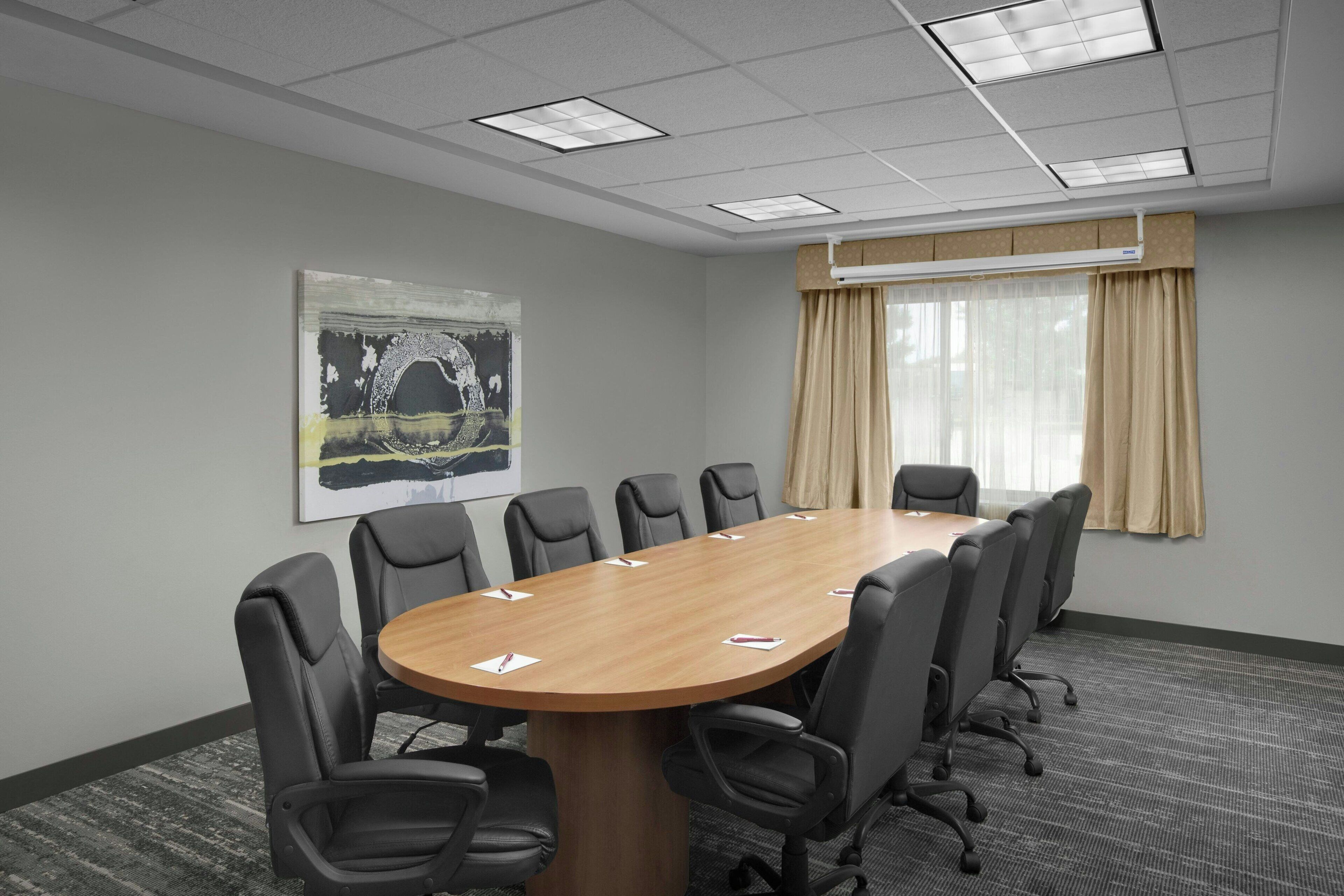 A conference room with a long table, black chairs, a large abstract painting, and a window with curtains.