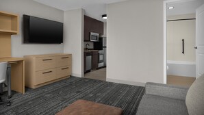 Une chambre d’hôtel moderne équipée d’une télévision à écran plat, d’un bureau avec une chaise, d’un canapé, d’un petit pouf et d’une kitchenette avec un four à micro-ondes et un lave-vaisselle.
