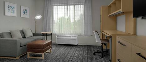 Een moderne hotelkamer met een grijze bank, een houten bureau, een flatscreen televisie en een groot raam met dunne gordijnen.
