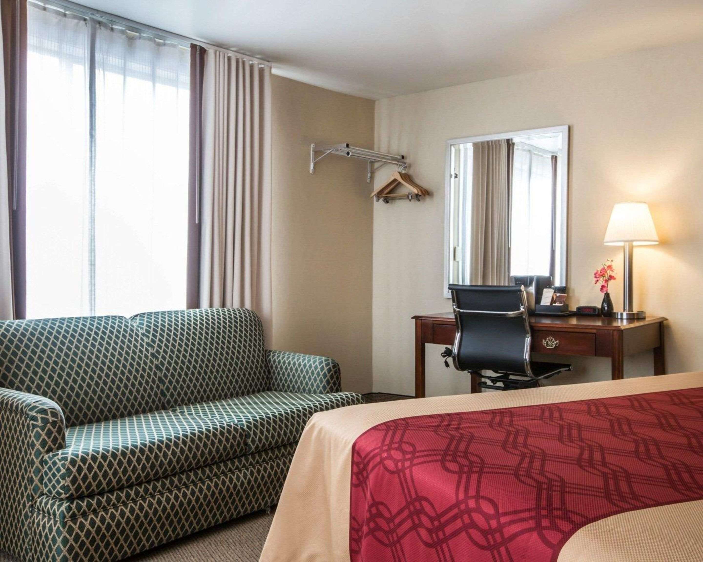 A hotel room with a patterned sofa, a desk with a chair, a mirror, and a lamp.