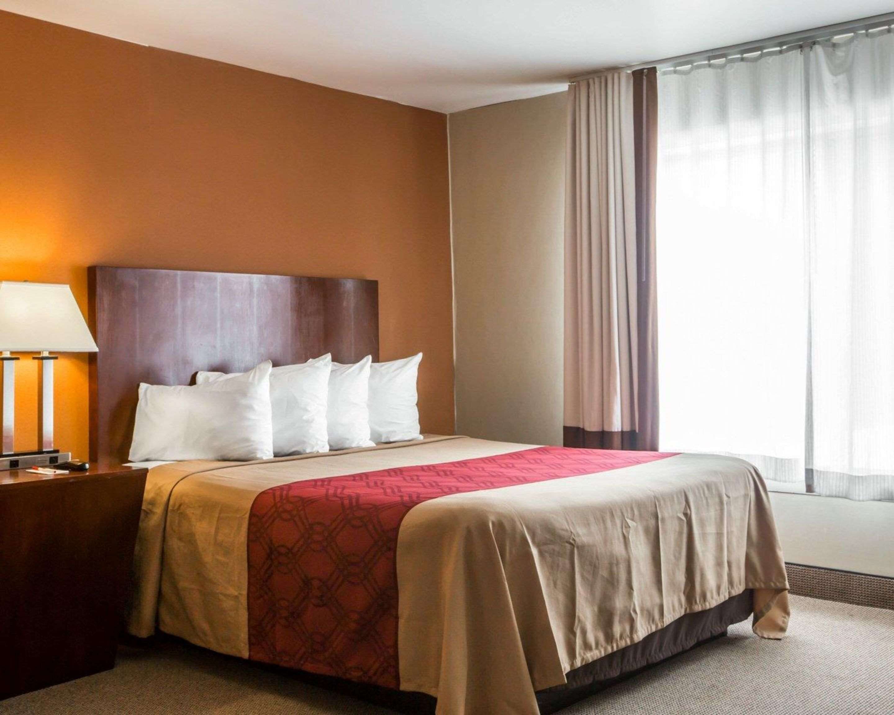 Habitación de hotel con una cama grande, cabecero de madera, mesita de noche con lámpara y ventana con cortinas.