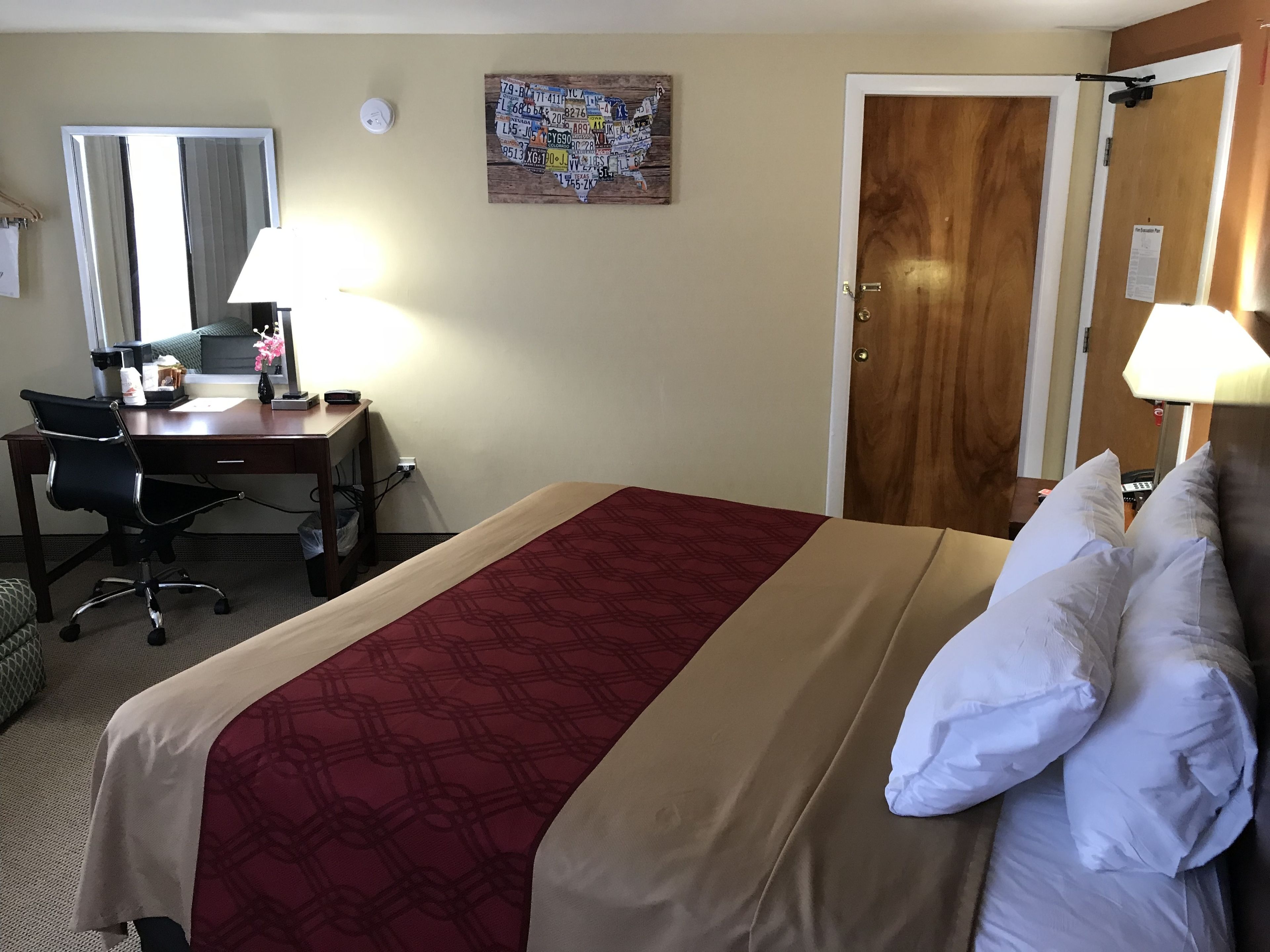 A hotel room with a bed, a desk, a chair, a lamp, a mirror, and a door.