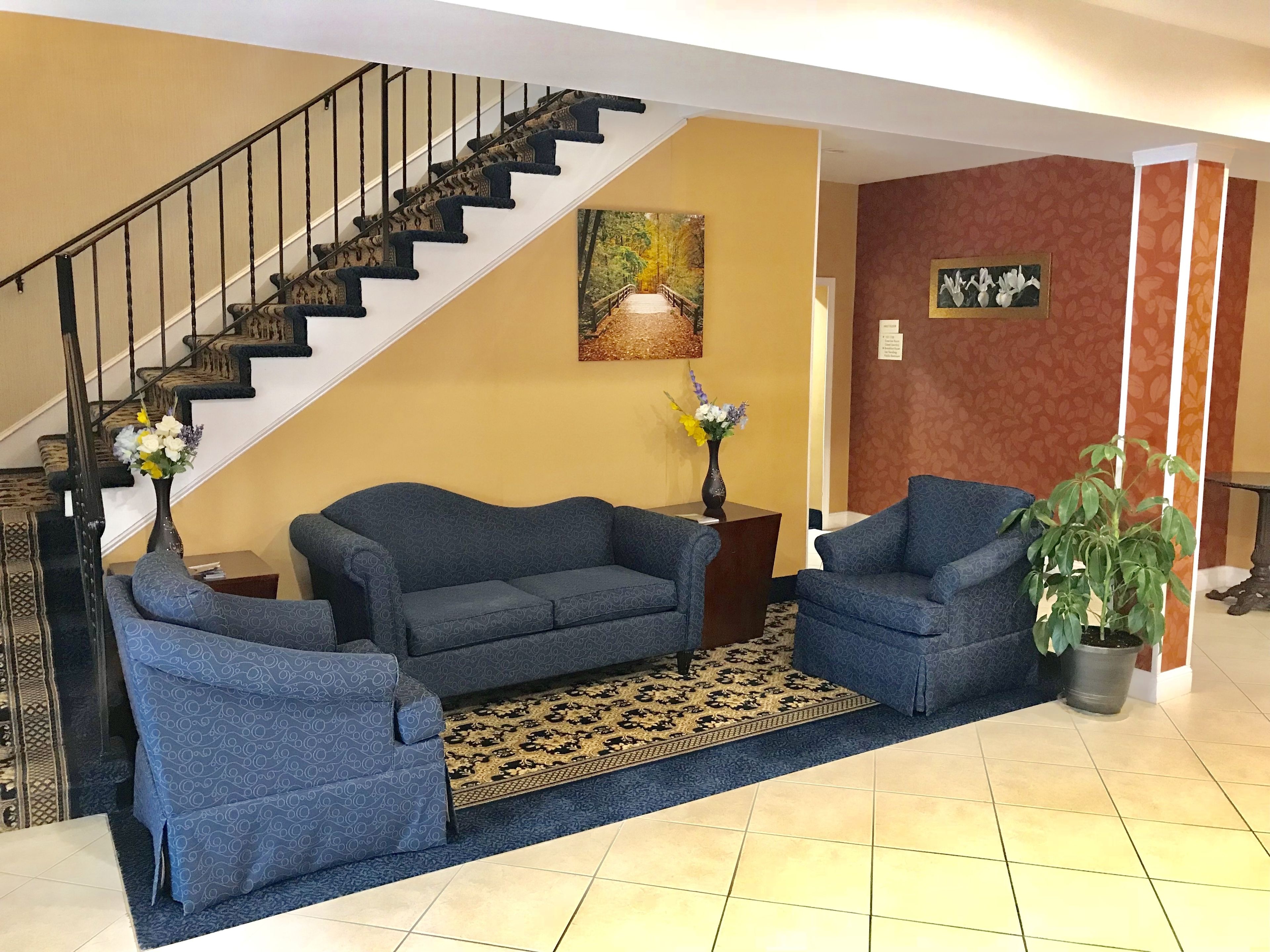 A living room with a staircase, two blue sofas, a patterned rug, a vase with flowers, and a potted plant.