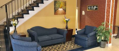 A living room with a staircase, two blue sofas, a patterned rug, a vase with flowers, and a potted plant.