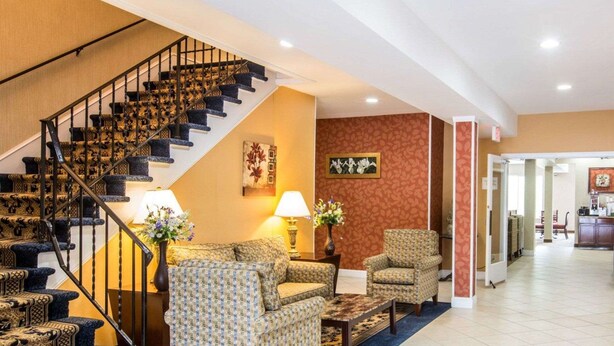 A hotel lobby with a staircase, patterned sofas, a coffee table, and a vase with flowers. - Econo Lodge Lake Region (West Ossipee)