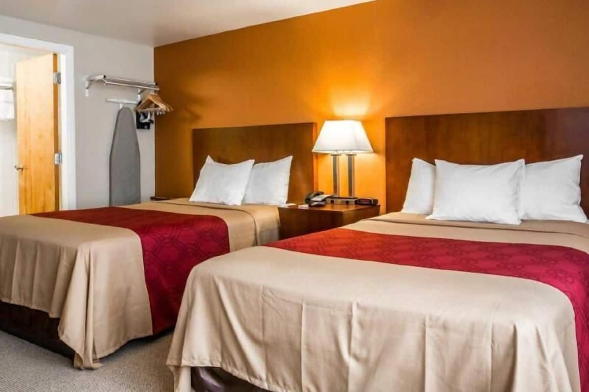 A hotel room with two beds, a nightstand with a lamp, a coat rack, and a door.