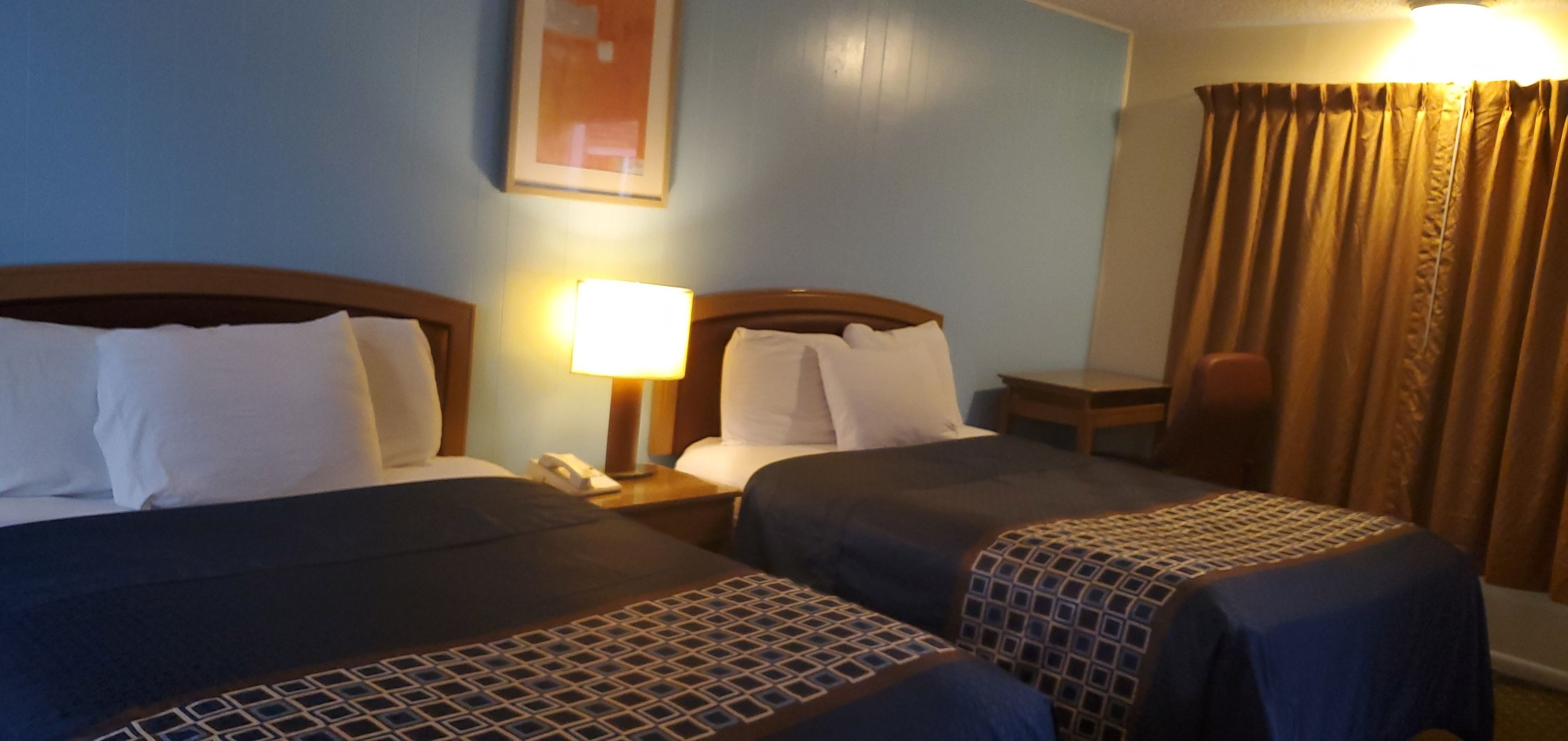 Habitación de hotel con dos camas, una mesita de noche con lámpara, una silla y un ventanal con cortinas.