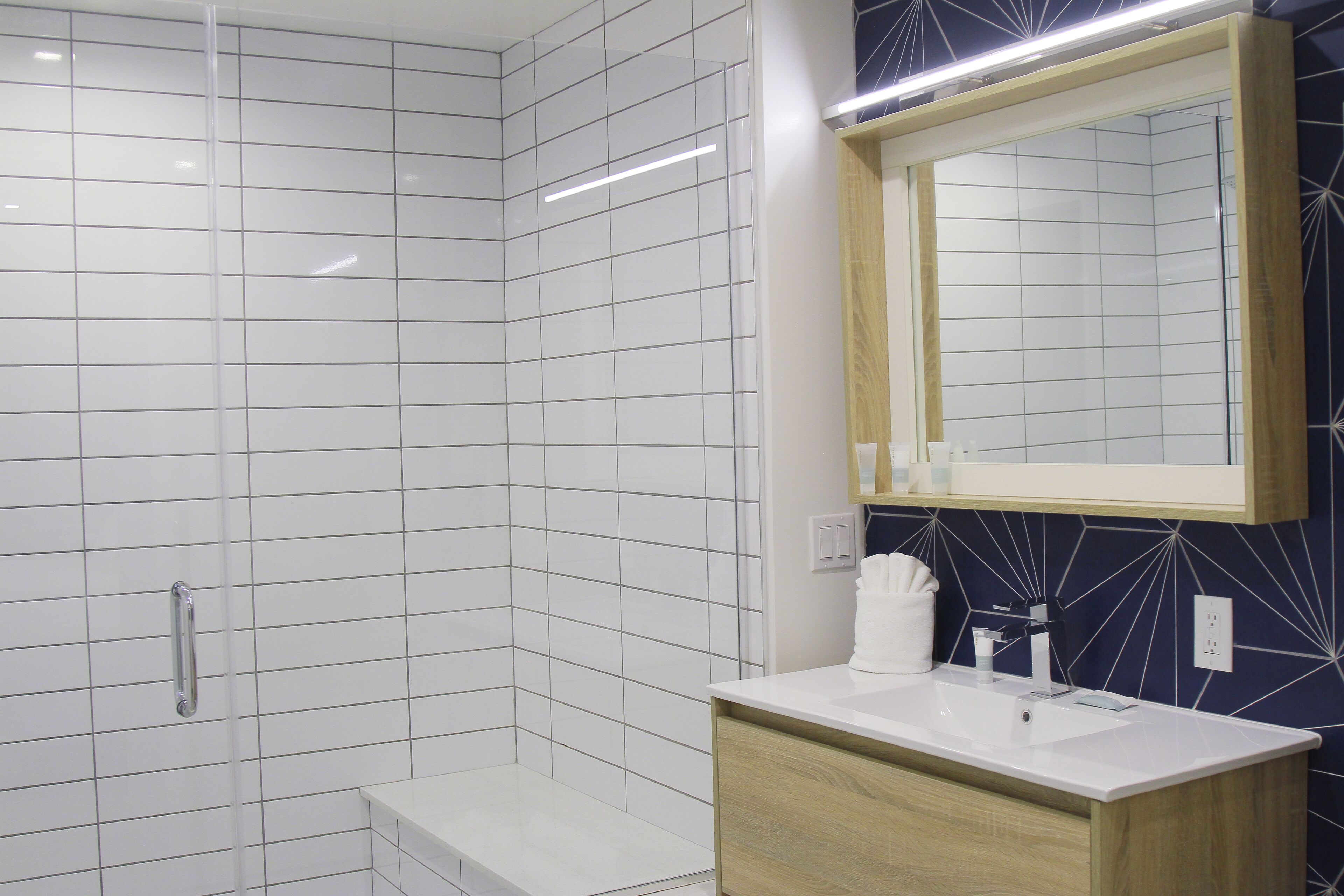 A modern bathroom with white tiled walls, a wooden vanity, a large mirror, and a shower area.