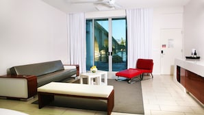Ein modernes Hotelzimmer mit grauem Sofa, einem roten Sessel, einem kleinen weißen Tisch mit Blumenvase und einer Bank.