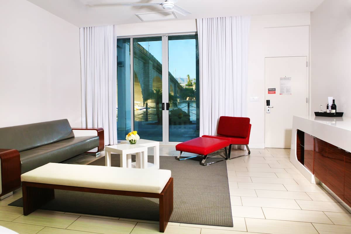 A modern hotel room with a grey sofa, a red armchair, a small white table with a vase of flowers, and a bench.
