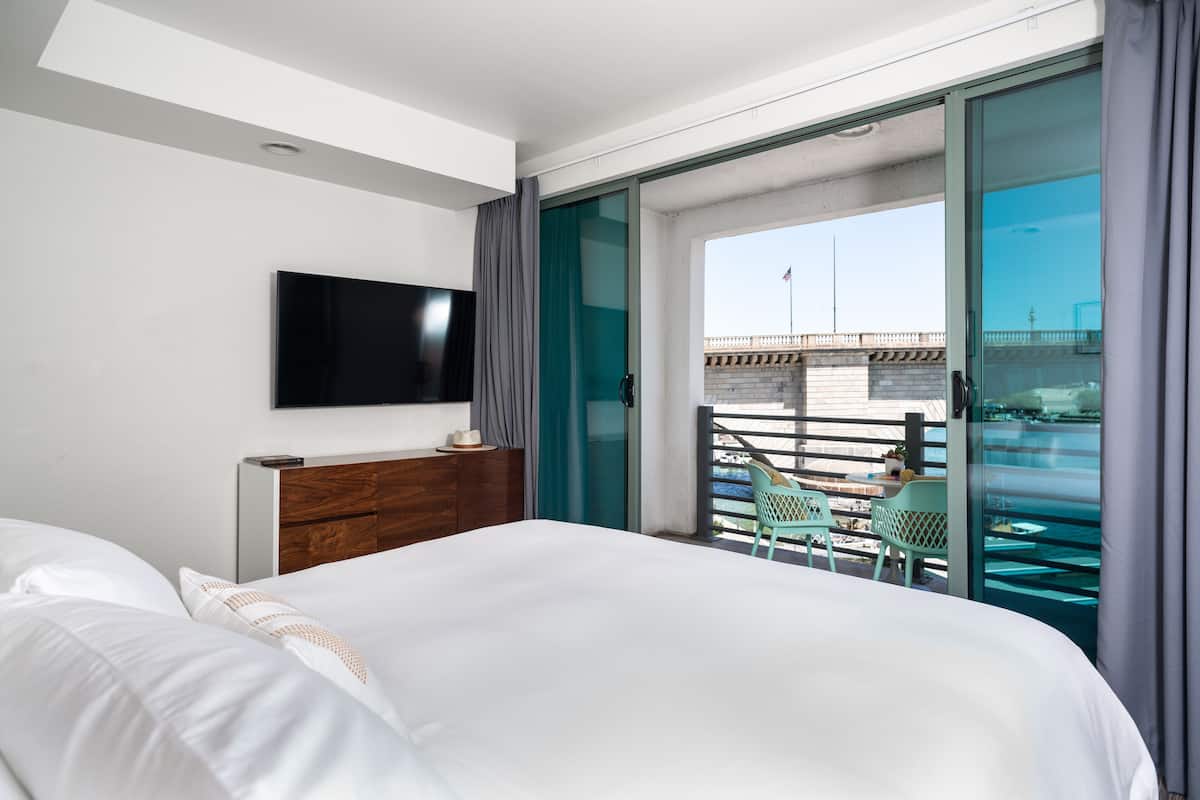 A hotel room with a large bed, a flat-screen TV mounted on the wall, a balcony with a view of the city, and a small table with chairs.
