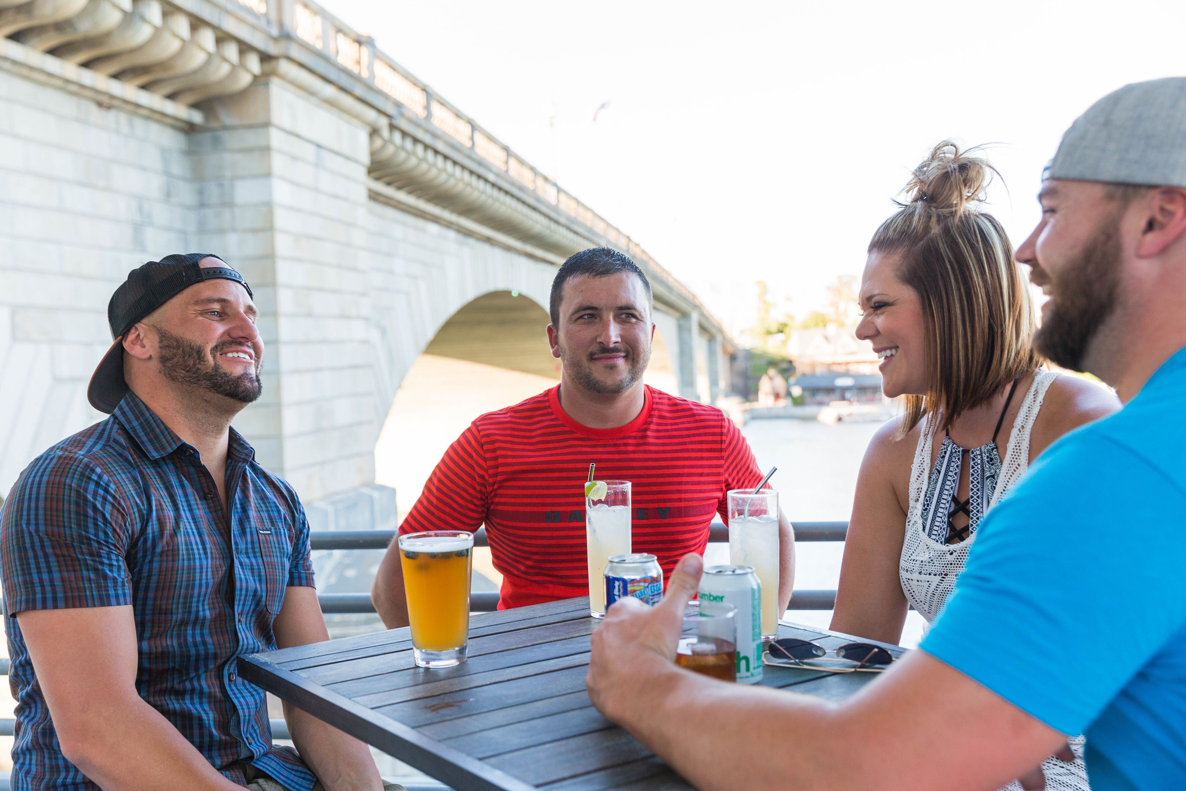 A group of people sitting at an outdoor table with drinks, a bridge in the background, and a casual dining setting.