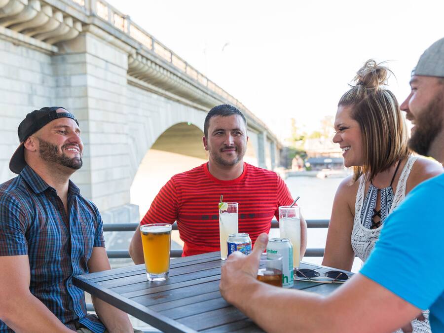 A group of people sitting at an outdoor table with drinks, a bridge in the background, and a casual dining setting.