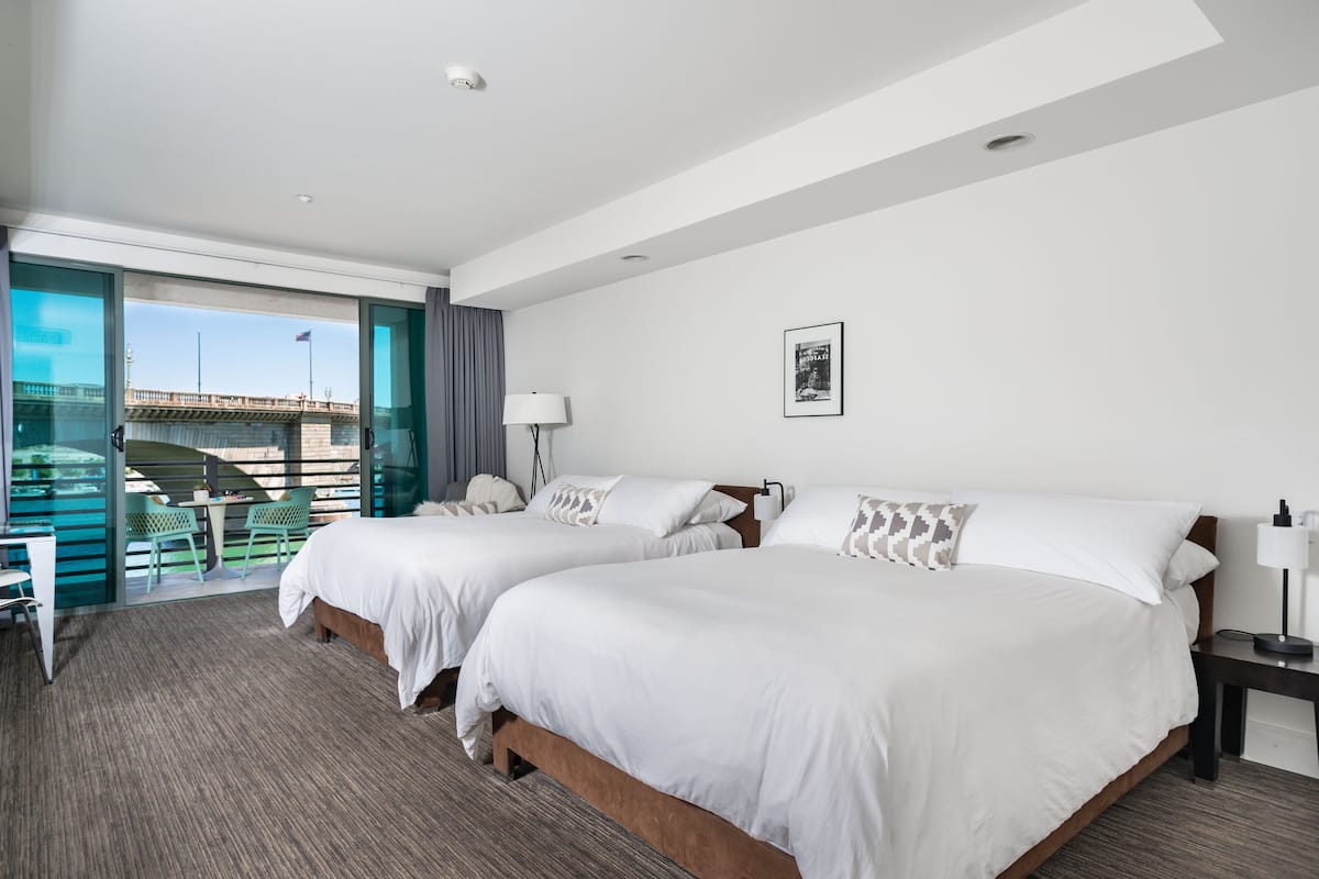 A hotel room with two beds, a desk, chairs, and a view of a bridge.
