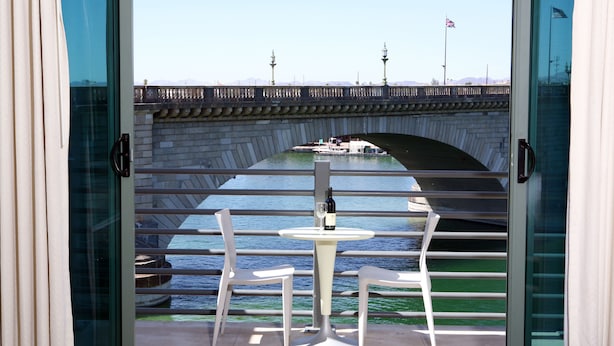 Blick von einem Balkon mit Tisch und Stühlen auf eine Brücke, an der ein Boot und Flaggen vorbeiführen.