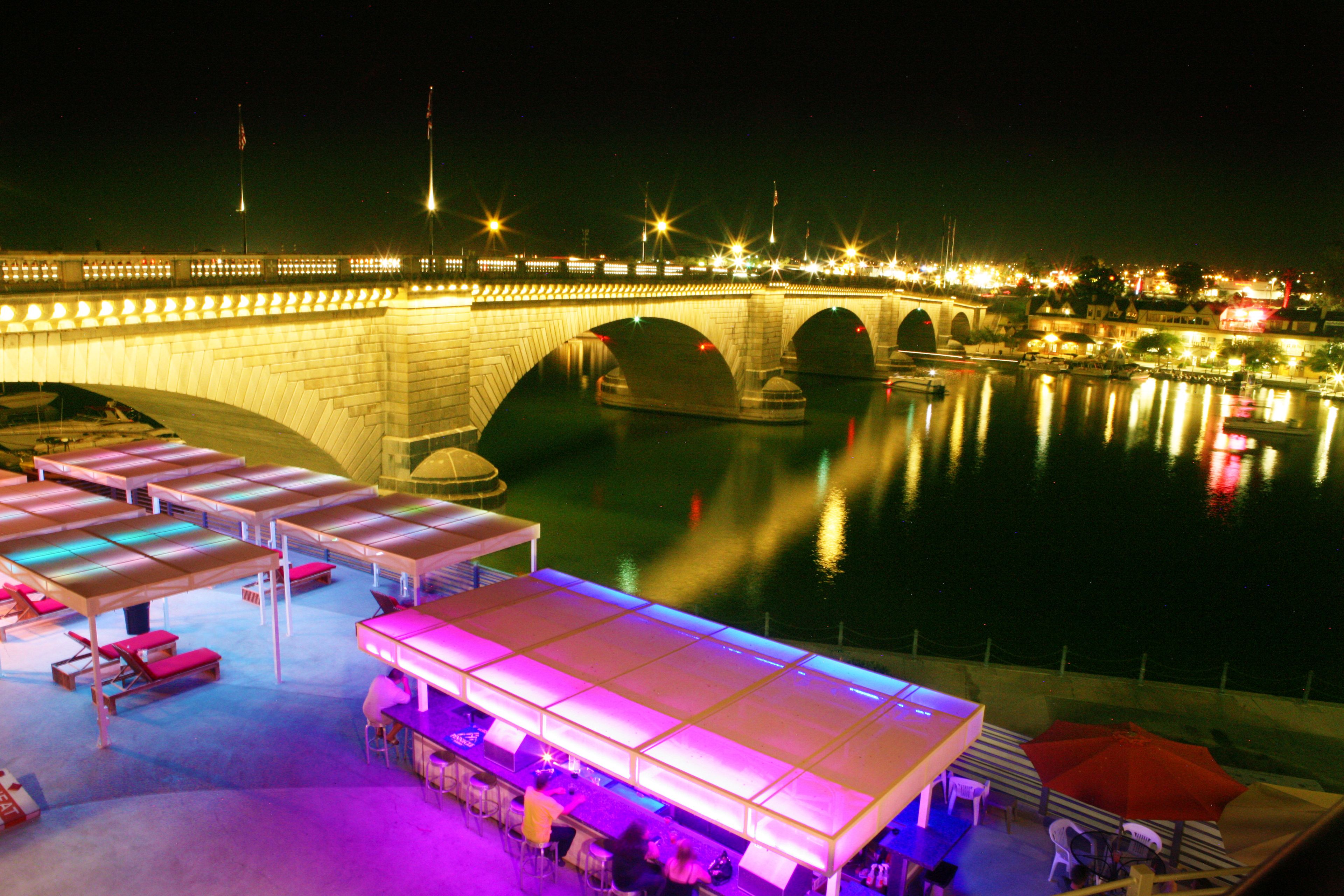 Un hotel frente al agua con zonas de estar al aire libre, un puente al fondo y una masa de agua tranquila que refleja las luces.