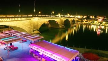 Un hotel frente al agua con zonas de estar al aire libre, un puente al fondo y una masa de agua tranquila que refleja las luces.