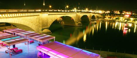 A waterfront hotel with outdoor seating areas, a bridge in the background, and a calm body of water reflecting the lights.