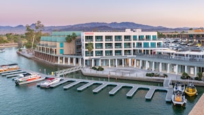 Ein modernes Hotel mit einer Marina, das sich durch ein mehrgeschossiges Gebäude mit großen Fenstern, eine Anlegestelle mit Booten und eine Aussicht auf Berge im Hintergrund auszeichnet.