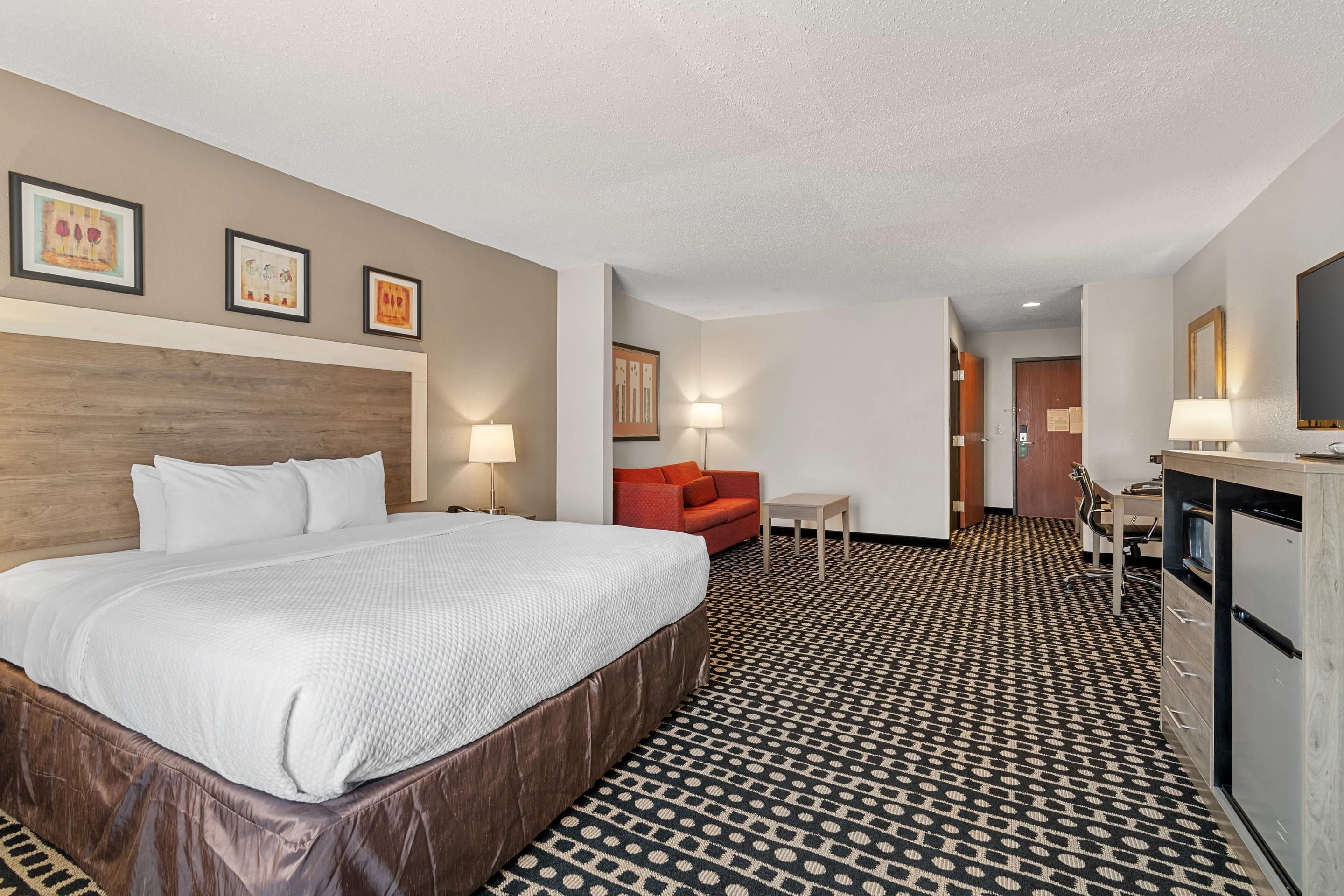 A hotel room with a large bed, a red sofa, a desk with a computer, and a kitchenette.