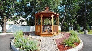 Un pabellón de madera con un banco, rodeado por un jardín con grava y acolchado rojo, y árboles al fondo.