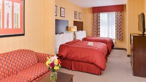 Habitación de hotel con sofá, dos camas, televisor y escritorio con un jarrón de flores.