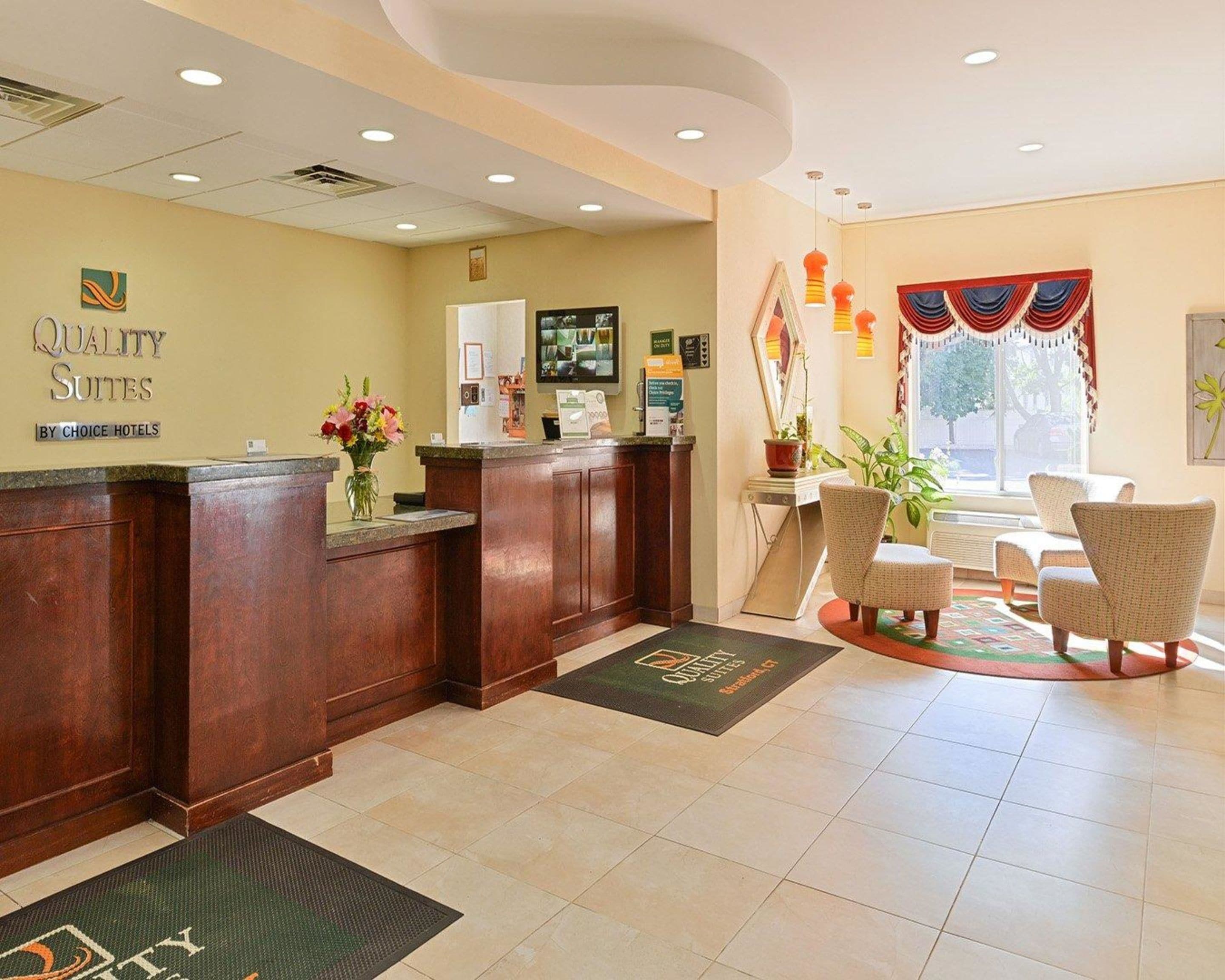 A hotel reception area with a wooden reception desk, a digital screen, and a seating area with chairs and a plant.