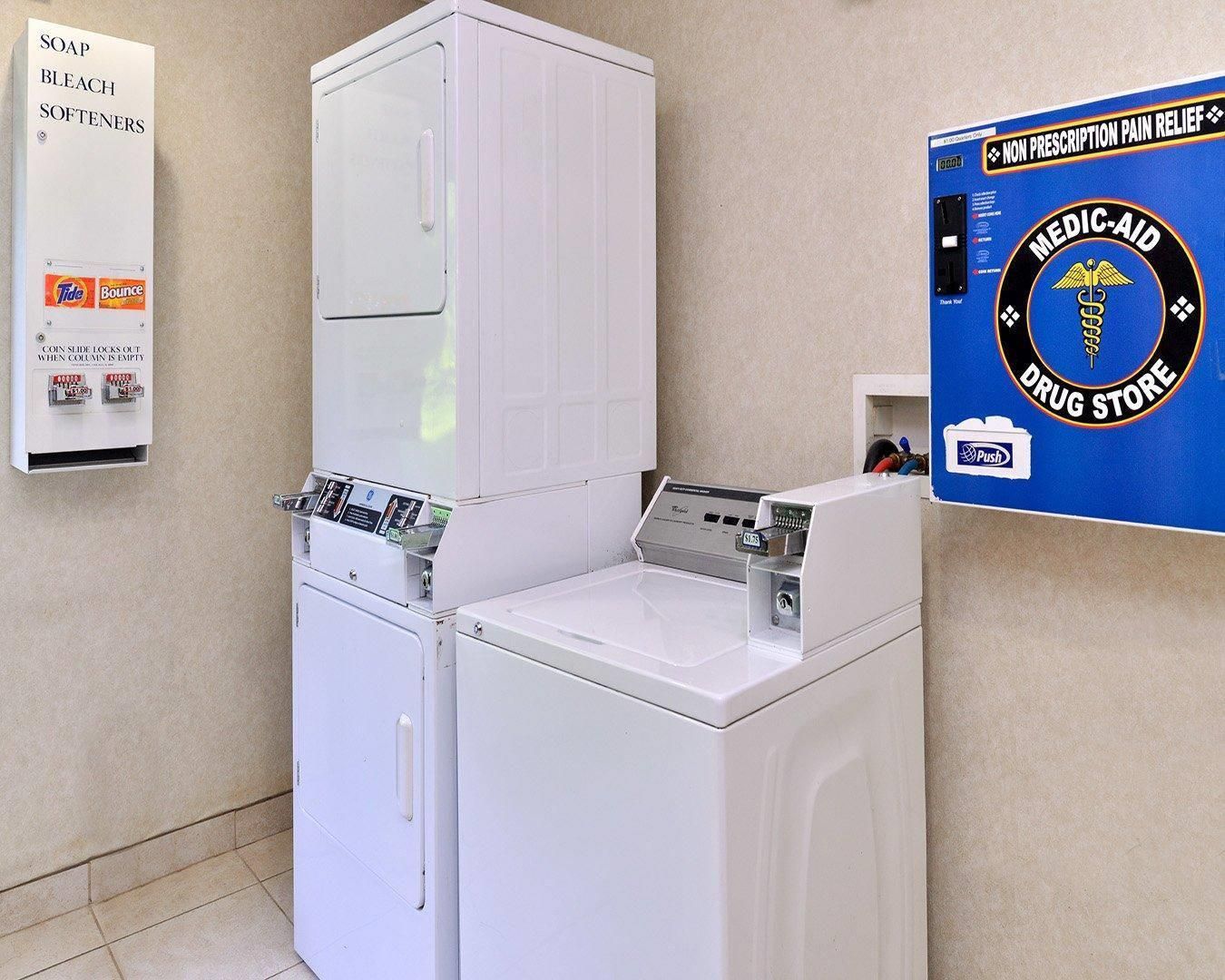 A laundromat with a coin-operated washer and dryer, a sign for a non-prescription pain relief drug store, and a sign for Tide and Bounce detergent.
