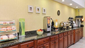 Zona de recepción de un hotel con mostrador que exhibe pasteles, un frutero, una máquina de café y un fabricante de gofres.