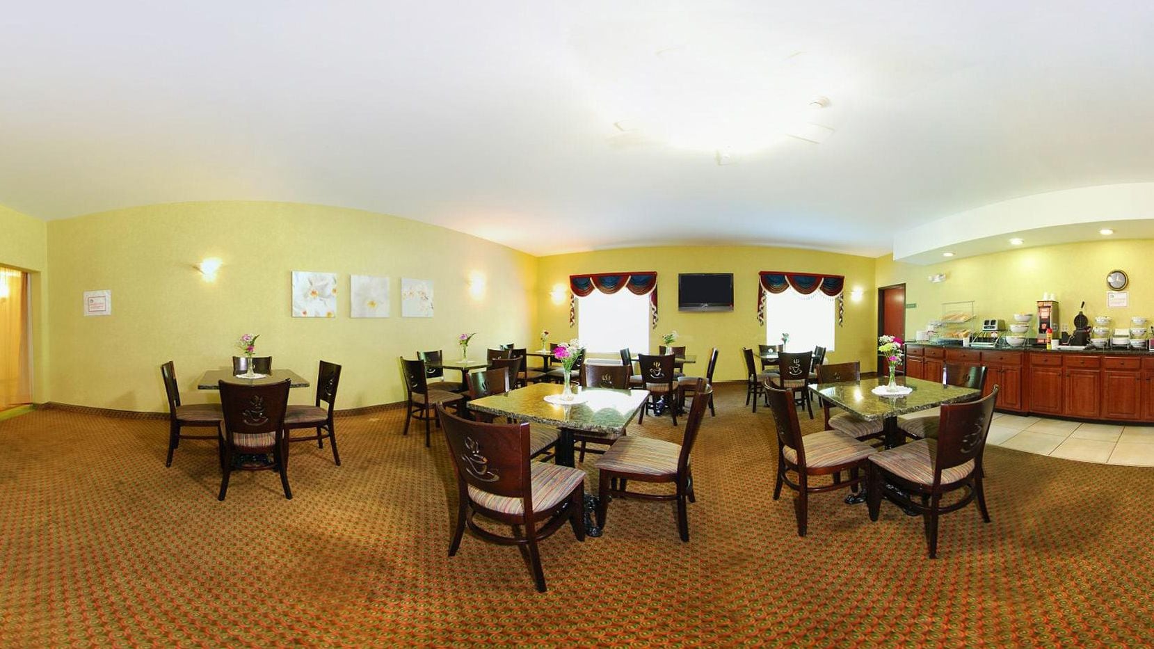 A dining area with round tables and chairs, a kitchen in the background, and a carpeted floor.