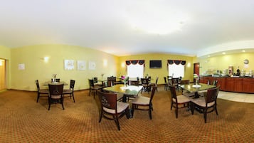 A dining area with round tables and chairs, a kitchen in the background, and a carpeted floor.