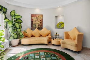 A living room with a sofa, armchairs, a coffee table, a large potted plant, a painting, and a decorative glass piece.