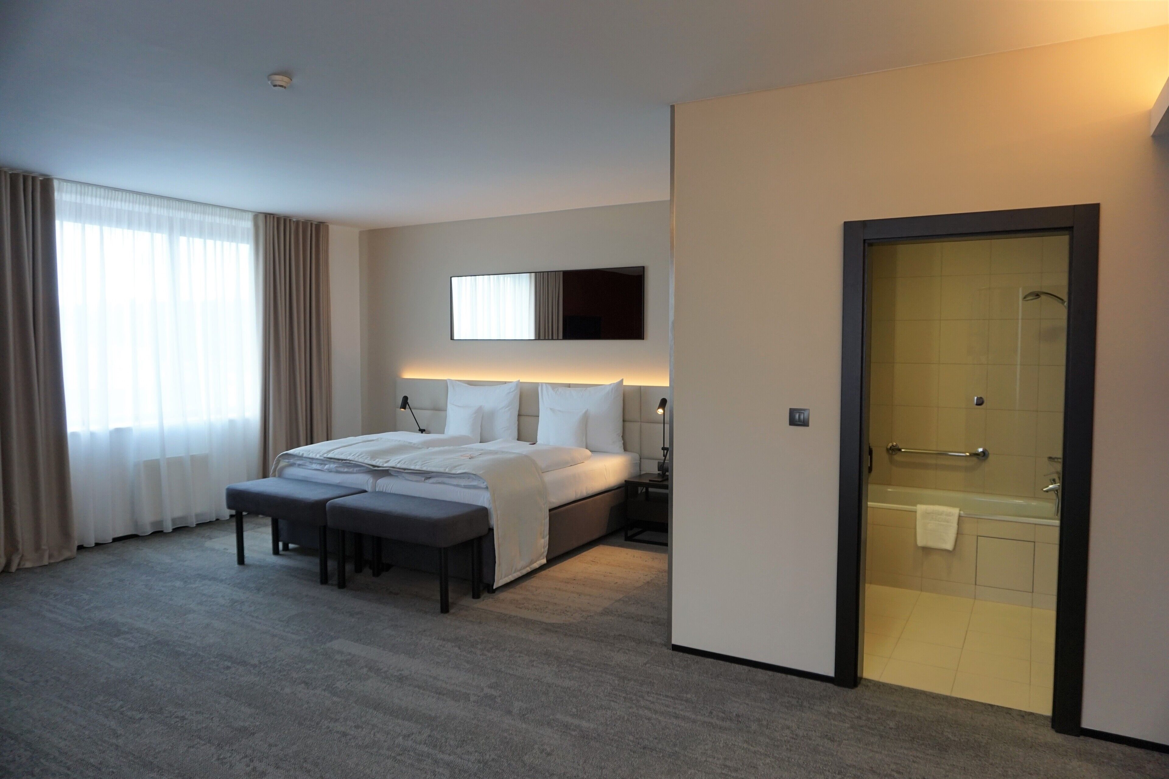 Habitación de hotel moderna con una cama grande, mesitas de noche, un banco, zona de ducha y un ventanal con cortinas.