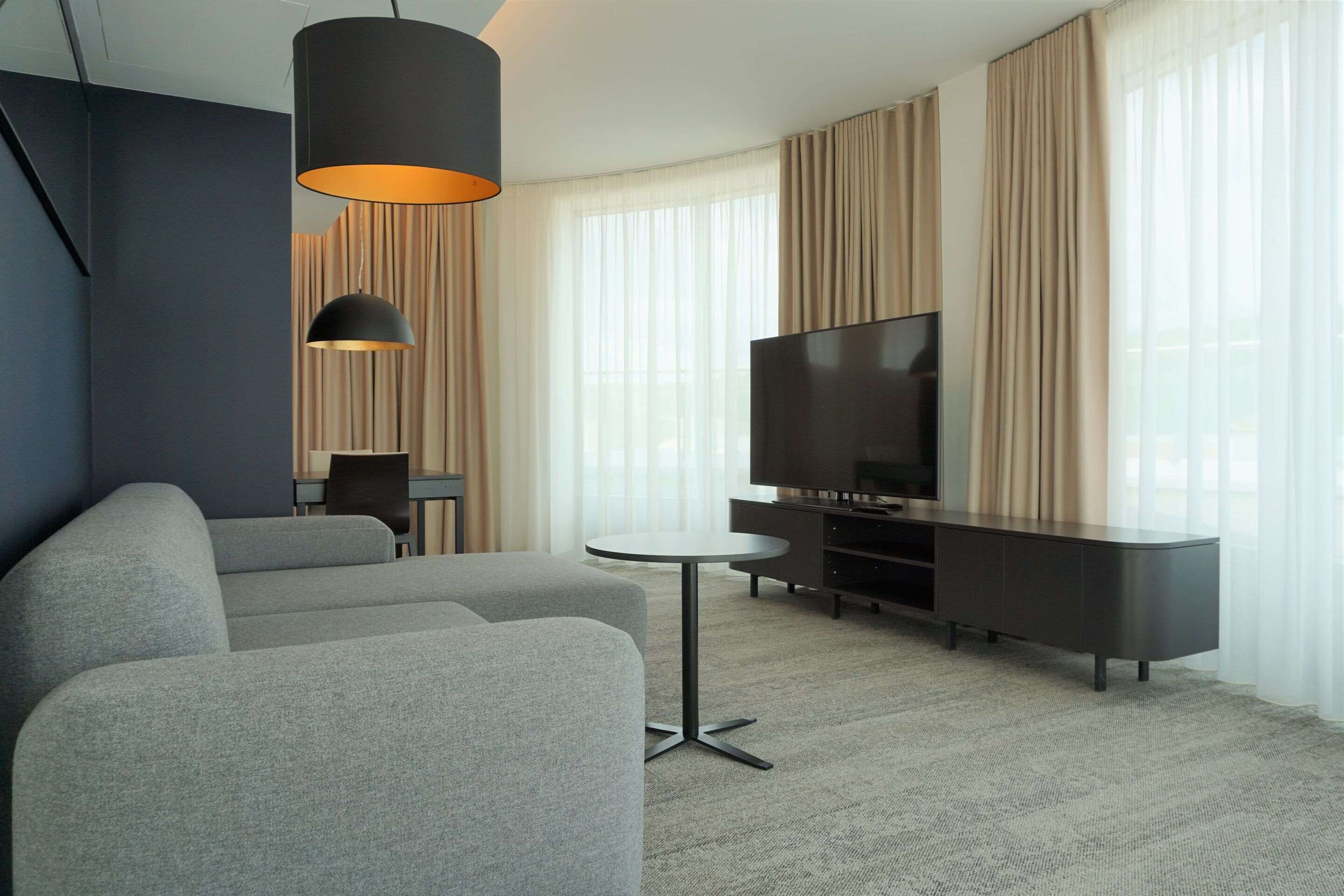 A modern living room with a grey sofa, a small round table, a TV on a black stand, and a large window with curtains.