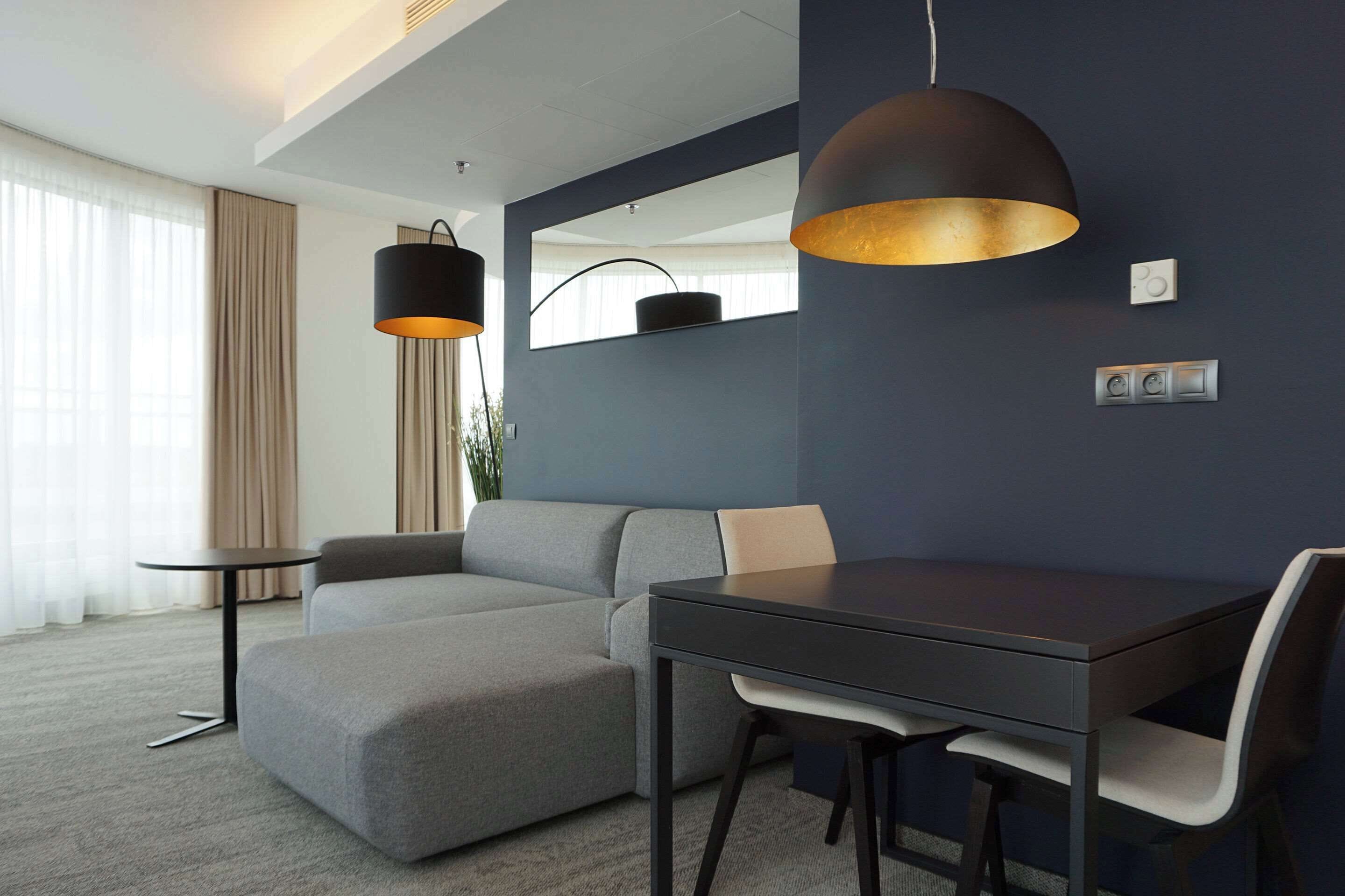 A modern hotel room with a grey sectional sofa, a small round table, a dark dining table with chairs, a large mirror, and a pendant light.
