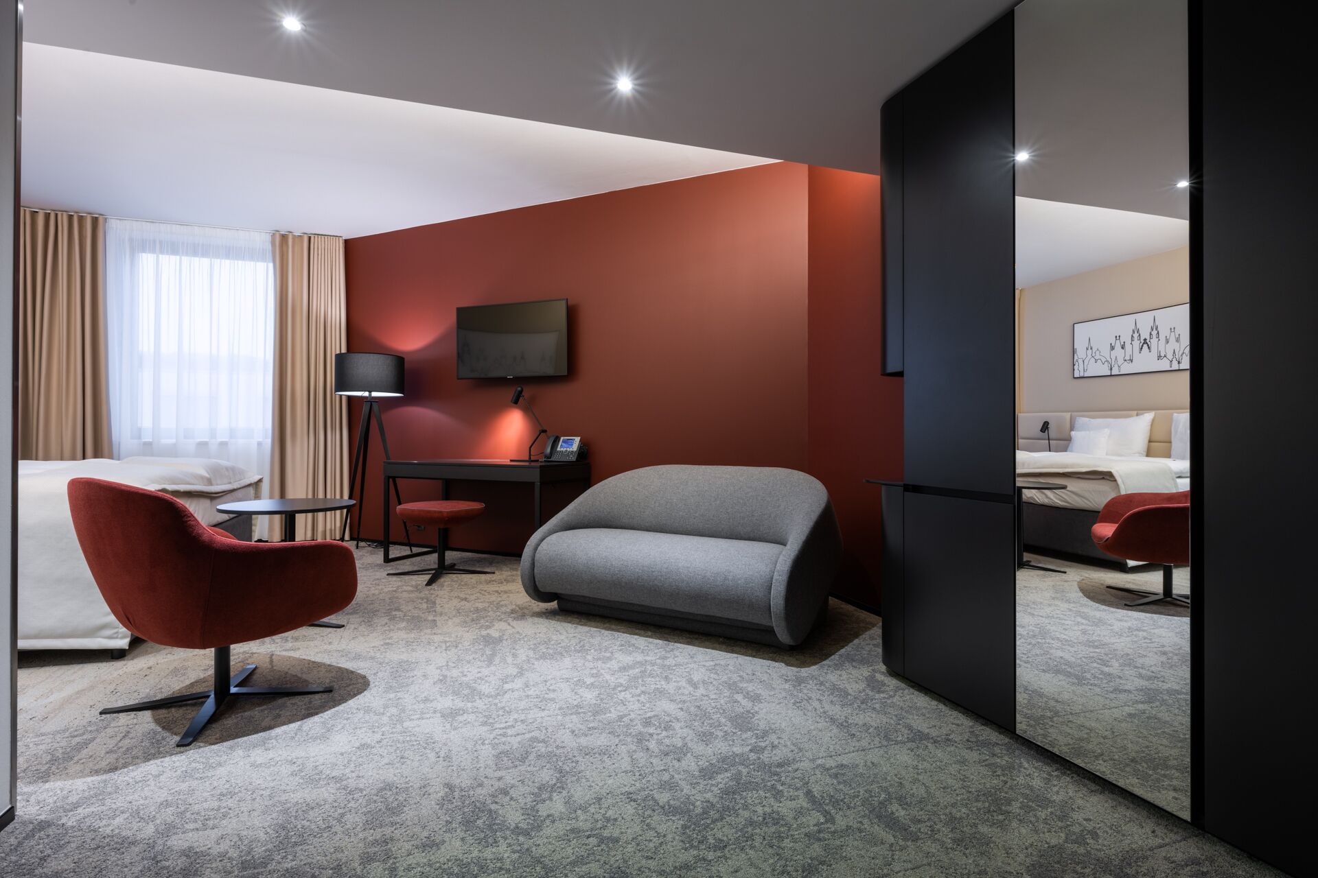 Une chambre d’hôtel moderne avec un canapé gris, un fauteuil rouge et un bureau avec une lampe.