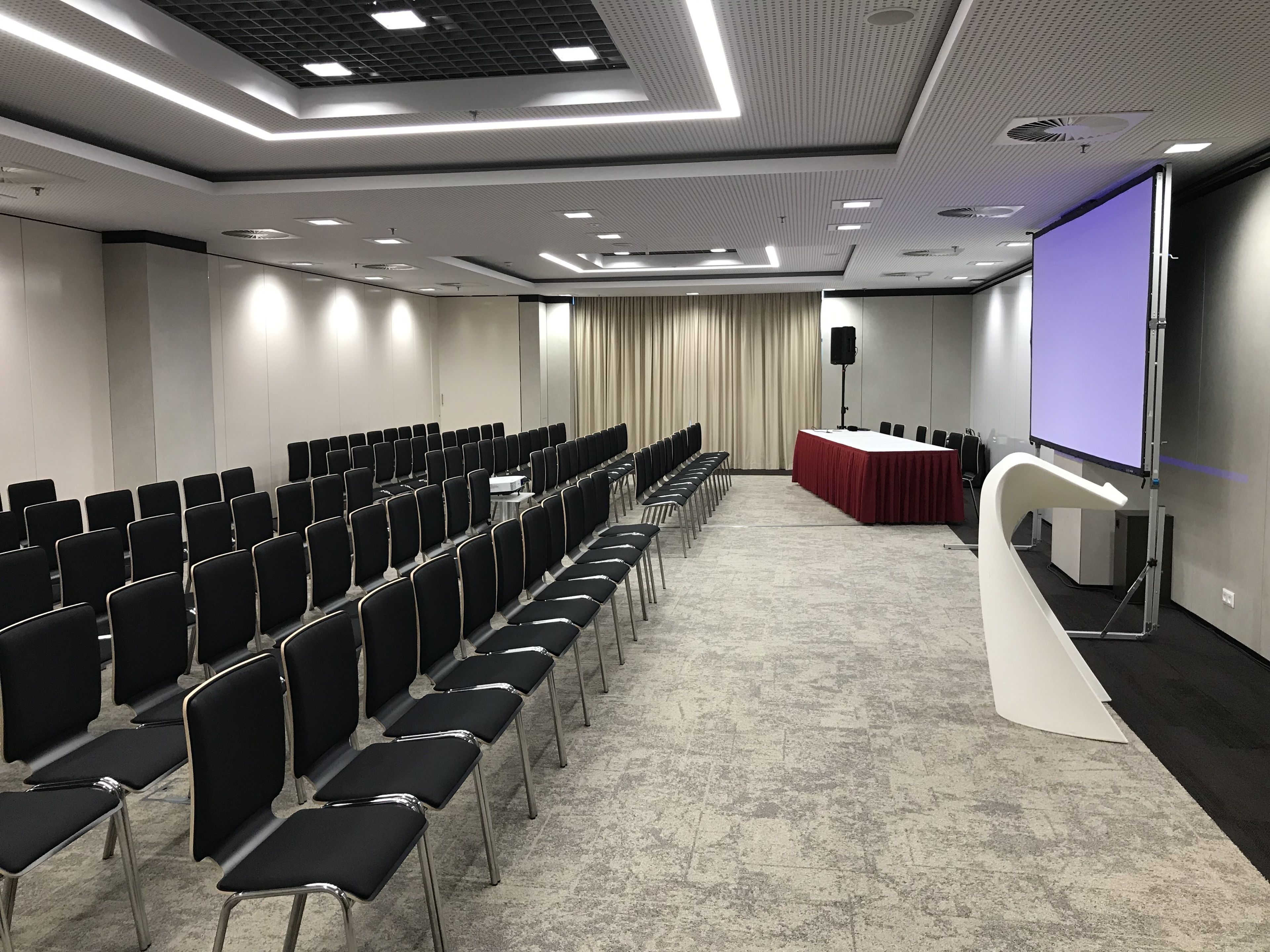 Un salón de conferencias con filas de sillas, un escenario con una mesa roja y una pantalla de proyector.