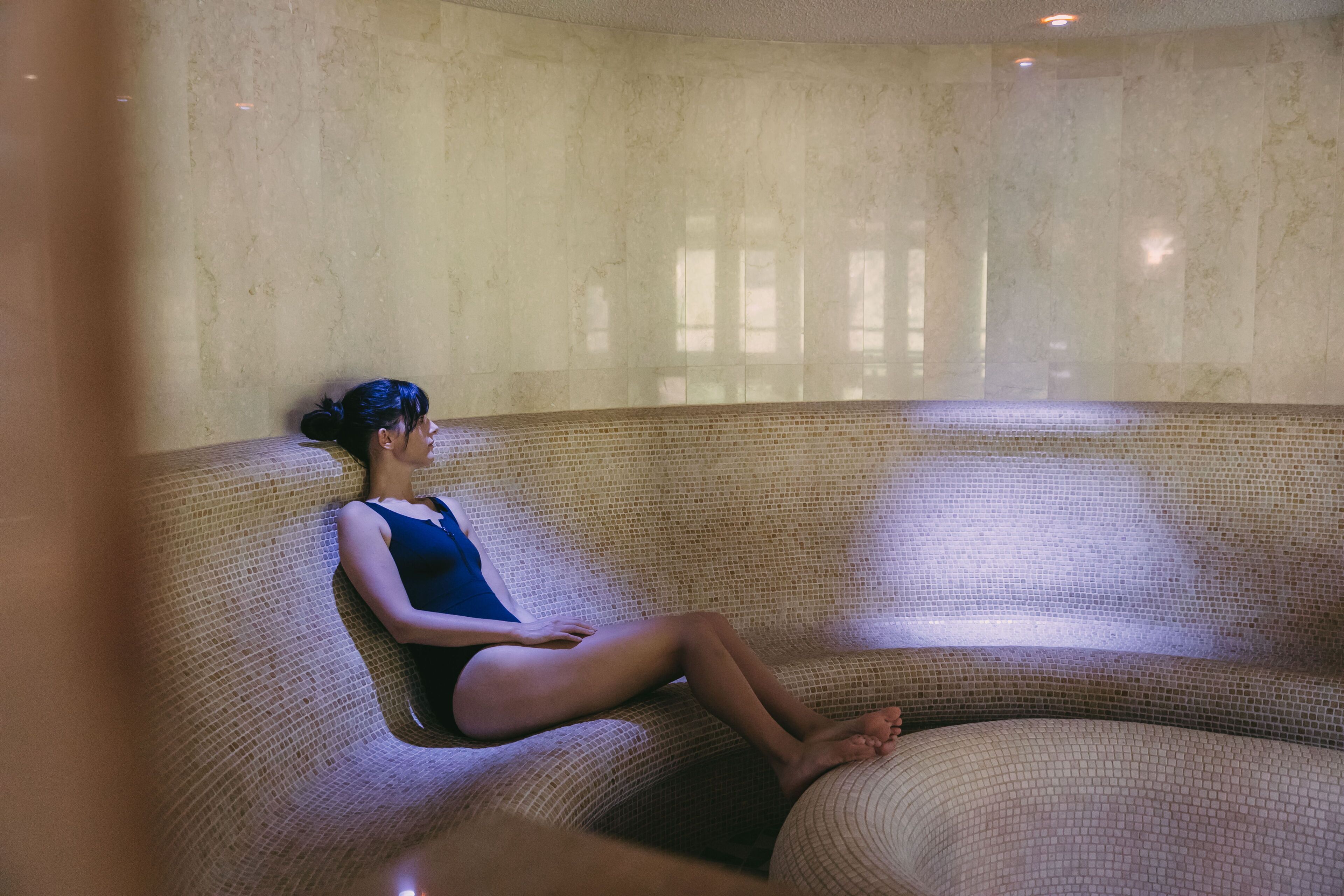 A person sitting on a textured, circular bench in a room with a plain wall and a window.