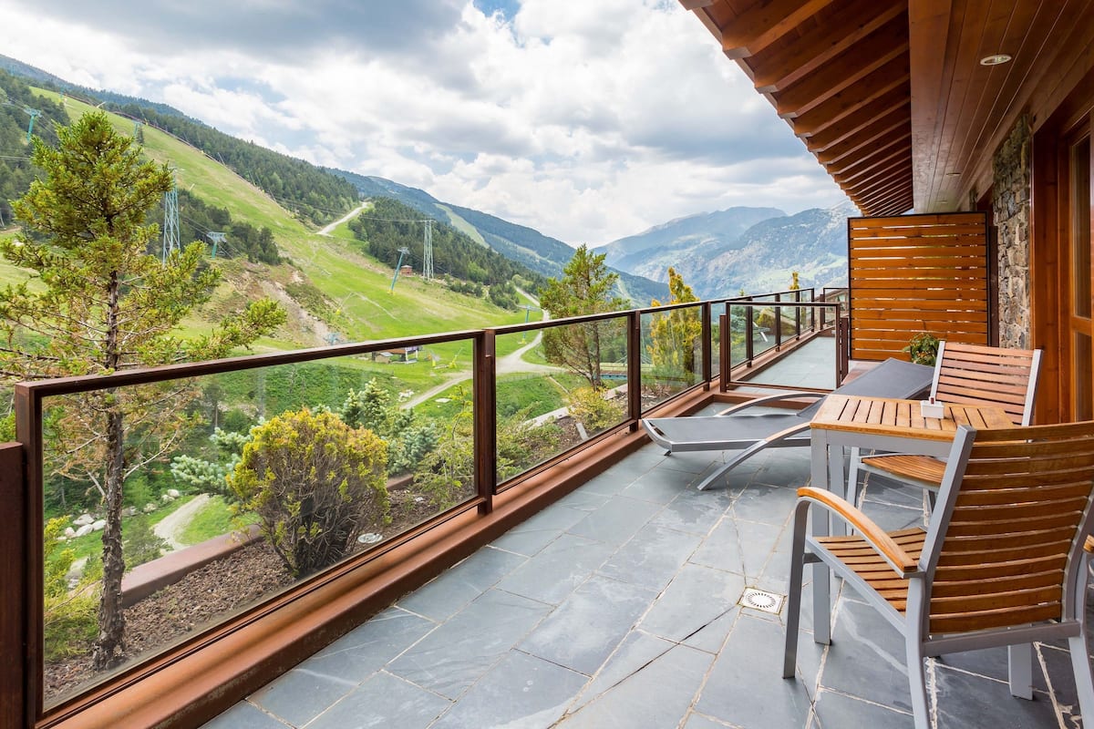 A balcony with a view of a mountainous landscape, a table with chairs, and stone walls.