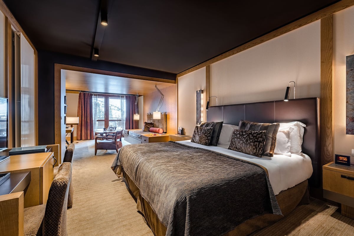 A modern hotel room with a large bed, a desk, and a chair. There is a window with curtains, a lamp, and a clock on the bedside table.