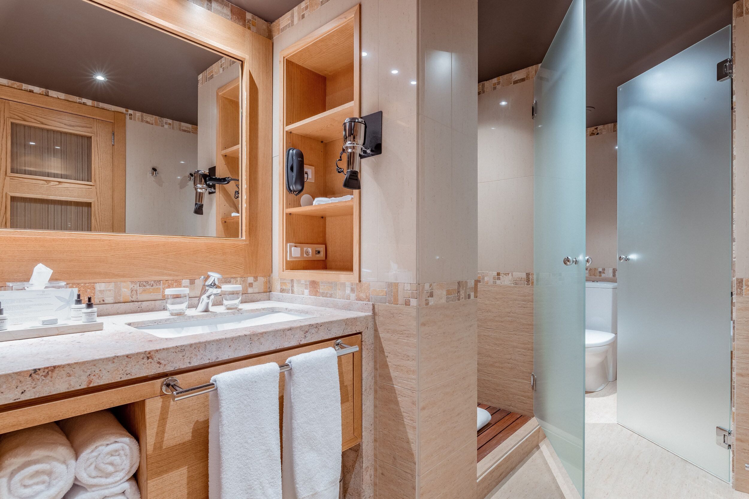 A modern bathroom with a large mirror, two sinks, a glass-enclosed shower, and wooden shelves with toiletries.