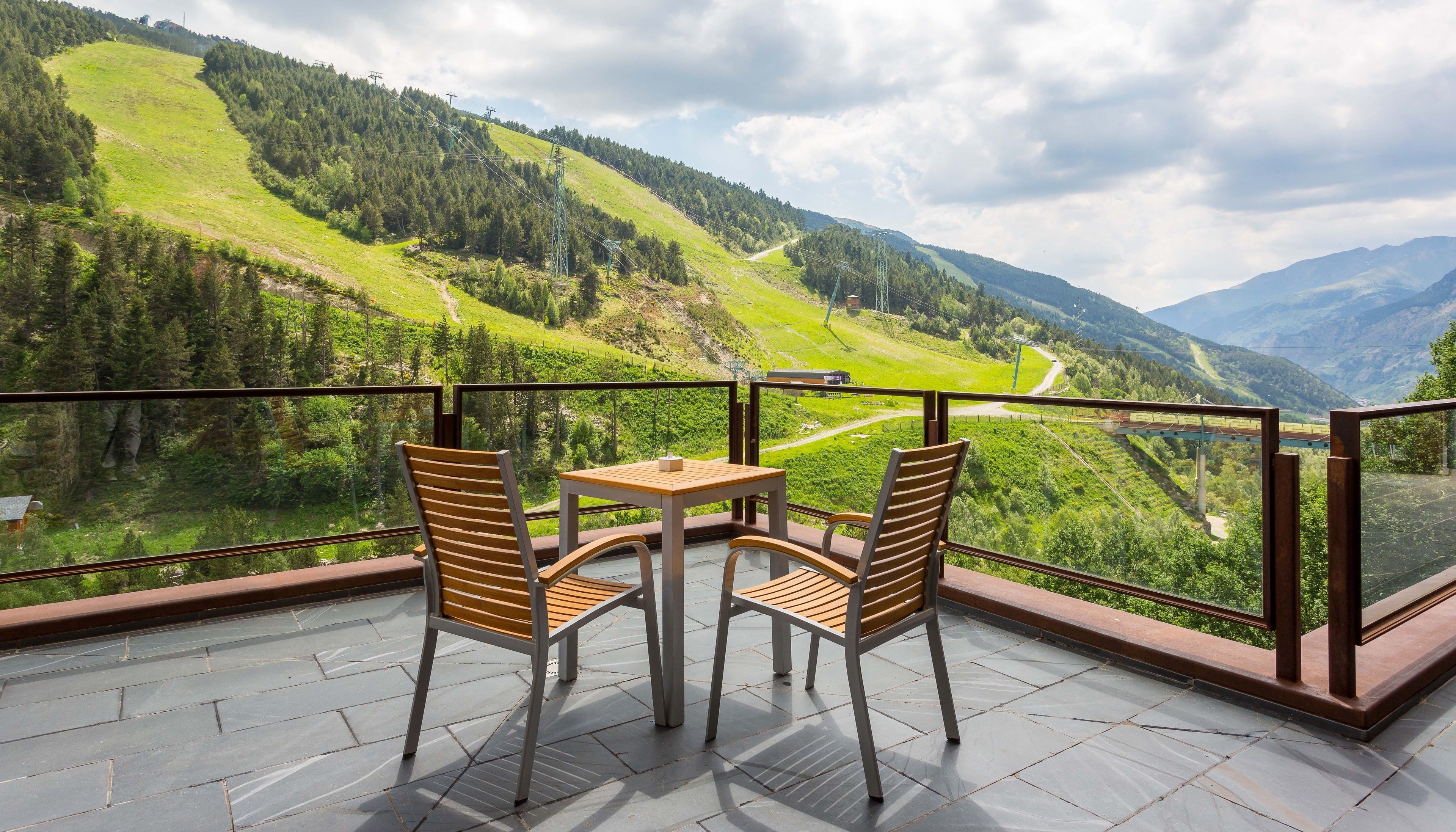 A balcony with two wooden chairs and a small table, overlooking a mountainous landscape with a clear sky.